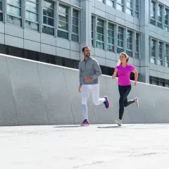 Ein Mann und eine Frau joggen nebeneinander. Ihre Atmung beim Joggen ist gleichmäßig, während sie an einer modernen Betonwand und einem Bürogebäude vorbeilaufen.