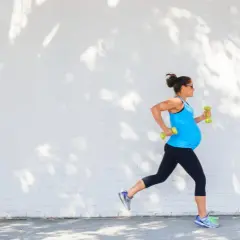 Eine schwangere Frau joggt mit Hanteln vor einer weißen Ziegelwand, beschattet von Baumblättern.