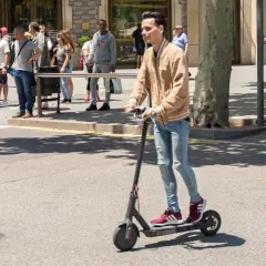 Ein junger Mann fährt mit einem E-Scooter im Ausland. Im Hintergrund warten Menschen auf den Bus und eine Frau fährt mit einem roten Leihfahrrad.