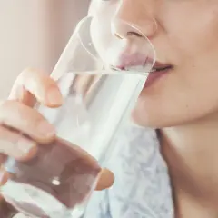 Frau trinkt nach dem Joggen Wasser. Sie nimmt das Glas mit beiden Händen und führt es zum Mund.