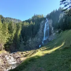 Wildstrubel Wasserfall: Ein majestätischer Anblick auf der UTMB-Route. Ein rauschender Wasserfall stürzt über Felsen in ein steiniges Flussbett, umgeben von dichten Nadelwäldern.