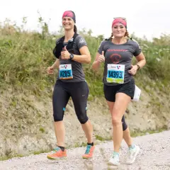 Zwei Läuferinnen auf einer Schotterpiste, die die Halbmarathon Distanz meistern. Sie lächeln und geben Daumen hoch, bereit für die Herausforderung.