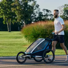 Ein Mann joggt mit Kinderwagen durch eine grüne Parklandschaft.
