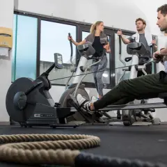 Ein Mann trainiert auf einem Rudergerät, während zwei weitere Personen auf Crosstrainern ausdauer trainieren.