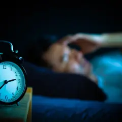 Ein Wecker zeigt 3 Uhr. Eine Person liegt im Bett, die Hand an der Stirn, und fragt sich: warum wache ich immer um 3 Uhr auf?