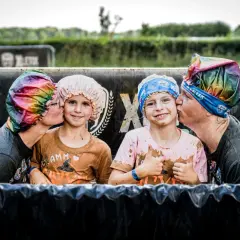 Zwei Erwachsene küssen zwei Kinder auf die Wange, alle tragen bunte Duschhauben. Ein fröhlicher Moment beim XLETIX Dresden Hindernislauf.