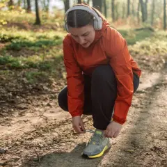 Frau mit Kopfhörern bindet ihre Laufschuhe auf einem Waldweg. Joggen im Alter ist eine tolle Möglichkeit, fit zu bleiben.