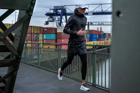 Mann joggt bei Regen über eine Brücke im Hafen. Container und Kräne im Hintergrund.