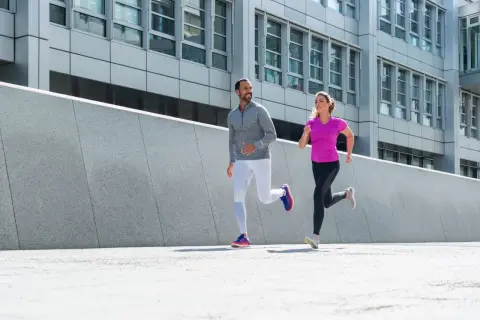 Ein Mann und eine Frau joggen nebeneinander. Ihre Atmung beim Joggen ist gleichmäßig, während sie an einer modernen Betonwand und einem Bürogebäude vorbeilaufen.
