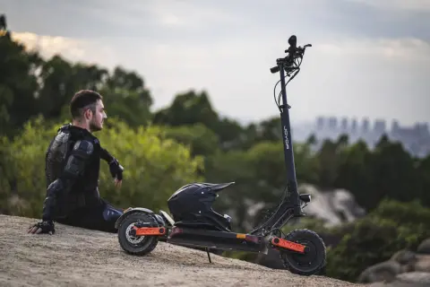 Mann in Schutzkleidung sitzt neben E-Roller mit Helm. Der e roller preis pro minute ist hier nicht ersichtlich, aber die Fahrt verspricht Abenteuer.