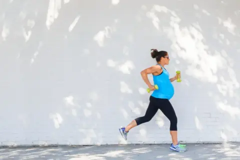 Eine schwangere Frau joggt mit Hanteln vor einer weißen Ziegelwand, beschattet von Baumblättern.