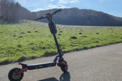 Ein alter E-Scooter steht am Straßenrand. Im Hintergrund weidet eine Schafherde auf einer grünen Wiese vor einem bewaldeten Hügel.