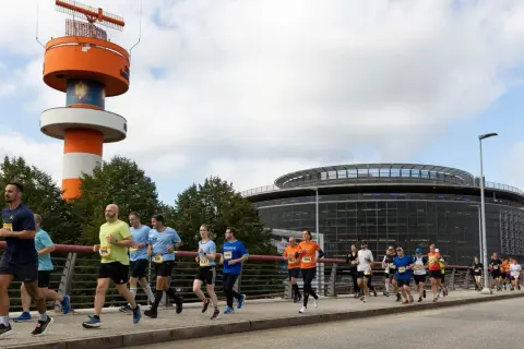 Läufer bei der Airport Race Hamburg, vorbei an einem markanten orange-weißen Turm und einem modernen Gebäude.