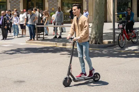 Ein junger Mann fährt mit einem E-Scooter im Ausland. Im Hintergrund warten Menschen auf den Bus und eine Frau fährt mit einem roten Leihfahrrad.