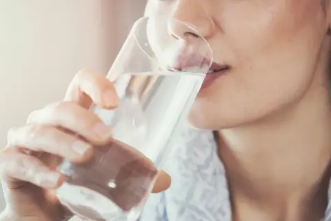 Frau trinkt nach dem Joggen Wasser. Sie nimmt das Glas mit beiden Händen und führt es zum Mund.
