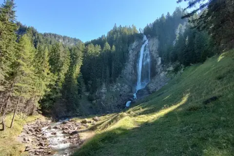 Wildstrubel Wasserfall: Ein majestätischer Anblick auf der UTMB-Route. Ein rauschender Wasserfall stürzt über Felsen in ein steiniges Flussbett, umgeben von dichten Nadelwäldern.
