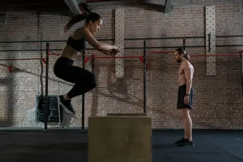 Frau springt auf eine Box, um Kondition aufzubauen. Mann beobachtet sie im Fitnessstudio.
