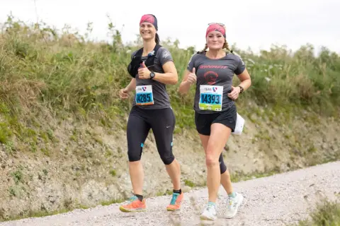 Zwei Läuferinnen auf einer Schotterpiste, die die Halbmarathon Distanz meistern. Sie lächeln und geben Daumen hoch, bereit für die Herausforderung.