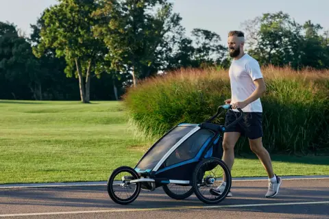 Ein Mann joggt mit Kinderwagen durch eine grüne Parklandschaft.