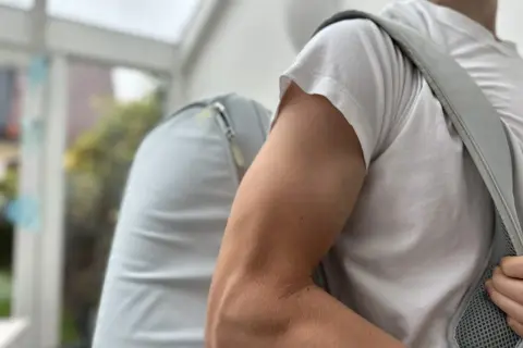 Mann mit Rucksack, der beim Anziehen der Jacke **Schmerzen im Oberarm** spürt.