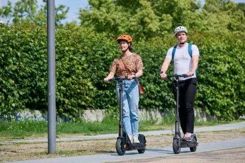 Mann und Frau fahren E-Scooter nebeneinander. Der Unterschied E-Roller und E-Scooter ist hier nicht ersichtlich, beide sind auf dem Weg.