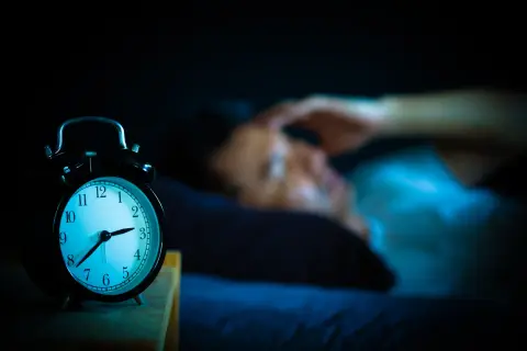 Ein Wecker zeigt 3 Uhr. Eine Person liegt im Bett, die Hand an der Stirn, und fragt sich: warum wache ich immer um 3 Uhr auf?