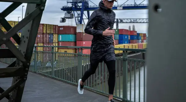 Mann joggt bei Regen über eine Brücke im Hafen. Container und Kräne im Hintergrund.