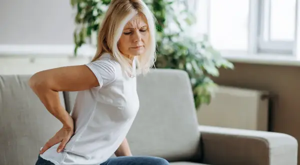 Frau mit schmerzen im lendenbereich sitzt auf einem Sofa und hält sich den Rücken.