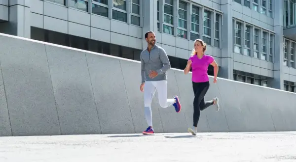 Ein Mann und eine Frau joggen nebeneinander. Ihre Atmung beim Joggen ist gleichmäßig, während sie an einer modernen Betonwand und einem Bürogebäude vorbeilaufen.
