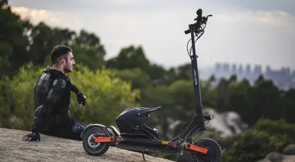 Mann in Schutzkleidung sitzt neben E-Roller mit Helm. Der e roller preis pro minute ist hier nicht ersichtlich, aber die Fahrt verspricht Abenteuer.