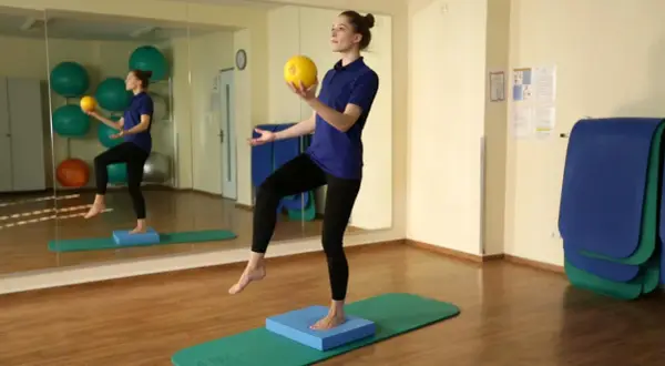 Eine Frau steht auf einem Bein auf einem blauen Kissen, während sie einen gelben Ball hält. Sie übt Balance.