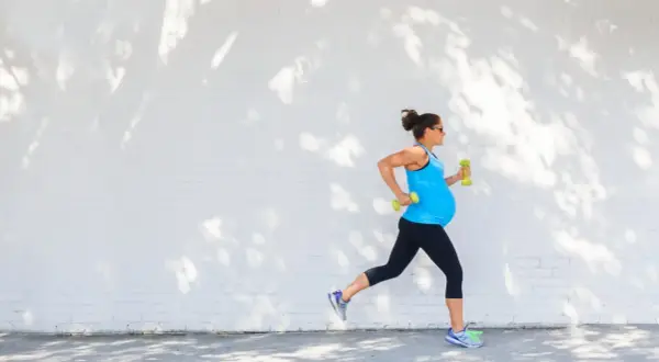 Eine schwangere Frau joggt mit Hanteln vor einer weißen Ziegelwand, beschattet von Baumblättern.