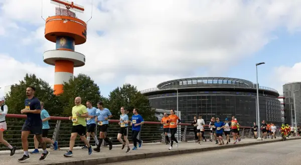 Läufer bei der Airport Race Hamburg, vorbei an einem markanten orange-weißen Turm und einem modernen Gebäude.