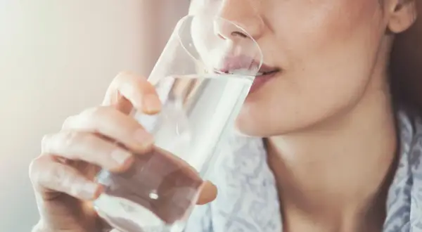 Frau trinkt nach dem Joggen Wasser. Sie nimmt das Glas mit beiden Händen und führt es zum Mund.