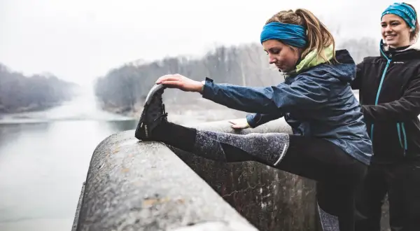 Zwei Frauen dehnen sich vor dem Laufen im Winter an einem Flussufer.