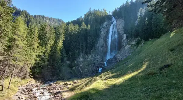 Wildstrubel Wasserfall: Ein majestätischer Anblick auf der UTMB-Route. Ein rauschender Wasserfall stürzt über Felsen in ein steiniges Flussbett, umgeben von dichten Nadelwäldern.