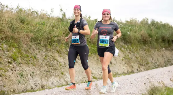 Zwei Läuferinnen auf einer Schotterpiste, die die Halbmarathon Distanz meistern. Sie lächeln und geben Daumen hoch, bereit für die Herausforderung.
