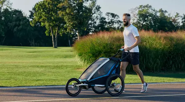 Ein Mann joggt mit Kinderwagen durch eine grüne Parklandschaft.