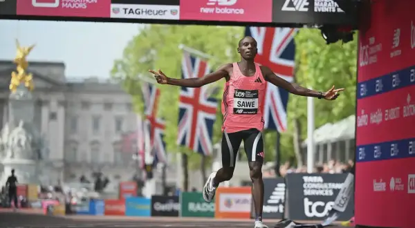 Läufer Sawe überquert die Ziellinie, vielleicht ein neuer weltrekord 5km. Britische Flaggen wehen im Hintergrund.
