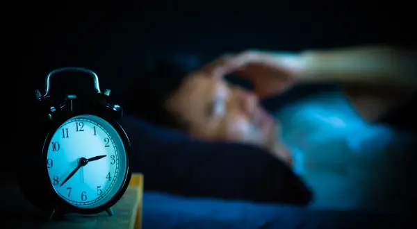 Ein Wecker zeigt 3 Uhr. Eine Person liegt im Bett, die Hand an der Stirn, und fragt sich: warum wache ich immer um 3 Uhr auf?