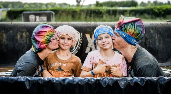 Zwei Erwachsene küssen zwei Kinder auf die Wange, alle tragen bunte Duschhauben. Ein fröhlicher Moment beim XLETIX Dresden Hindernislauf.