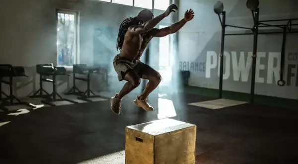 Mann macht explosive Box Jumps in einem Fitnessstudio. Rauch und Lichteffekte unterstreichen die Dynamik.