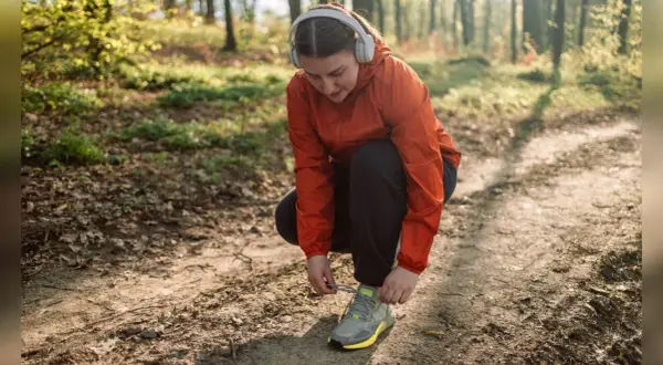 Frau mit Kopfhörern bindet ihre Laufschuhe auf einem Waldweg. Joggen im Alter ist eine tolle Möglichkeit, fit zu bleiben.