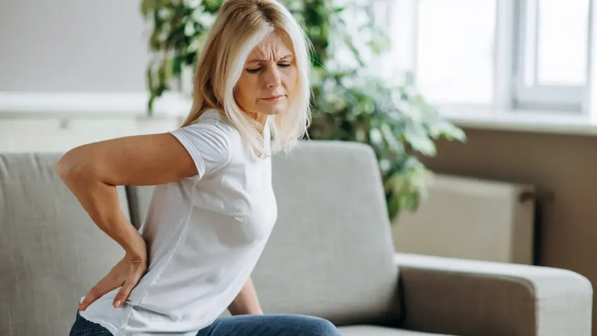Frau mit schmerzen im lendenbereich sitzt auf einem Sofa und hält sich den Rücken.