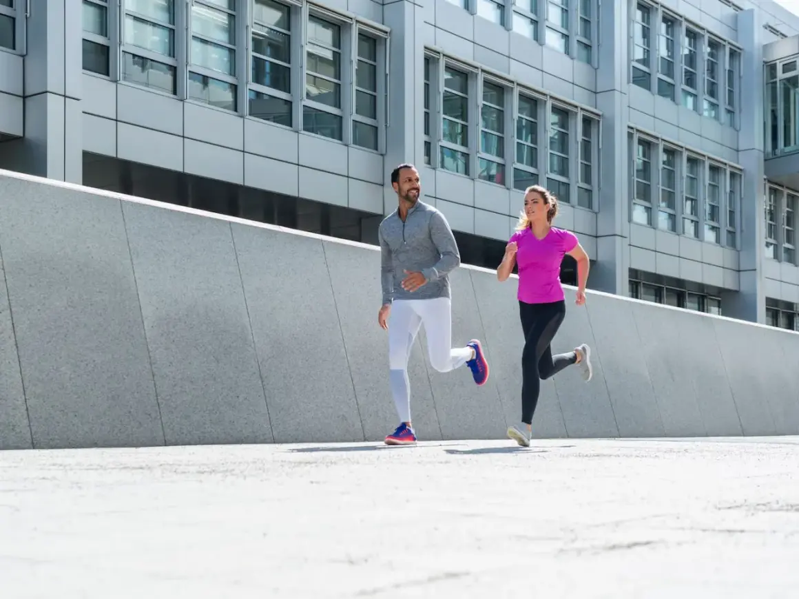 Ein Mann und eine Frau joggen nebeneinander. Ihre Atmung beim Joggen ist gleichmäßig, während sie an einer modernen Betonwand und einem Bürogebäude vorbeilaufen.