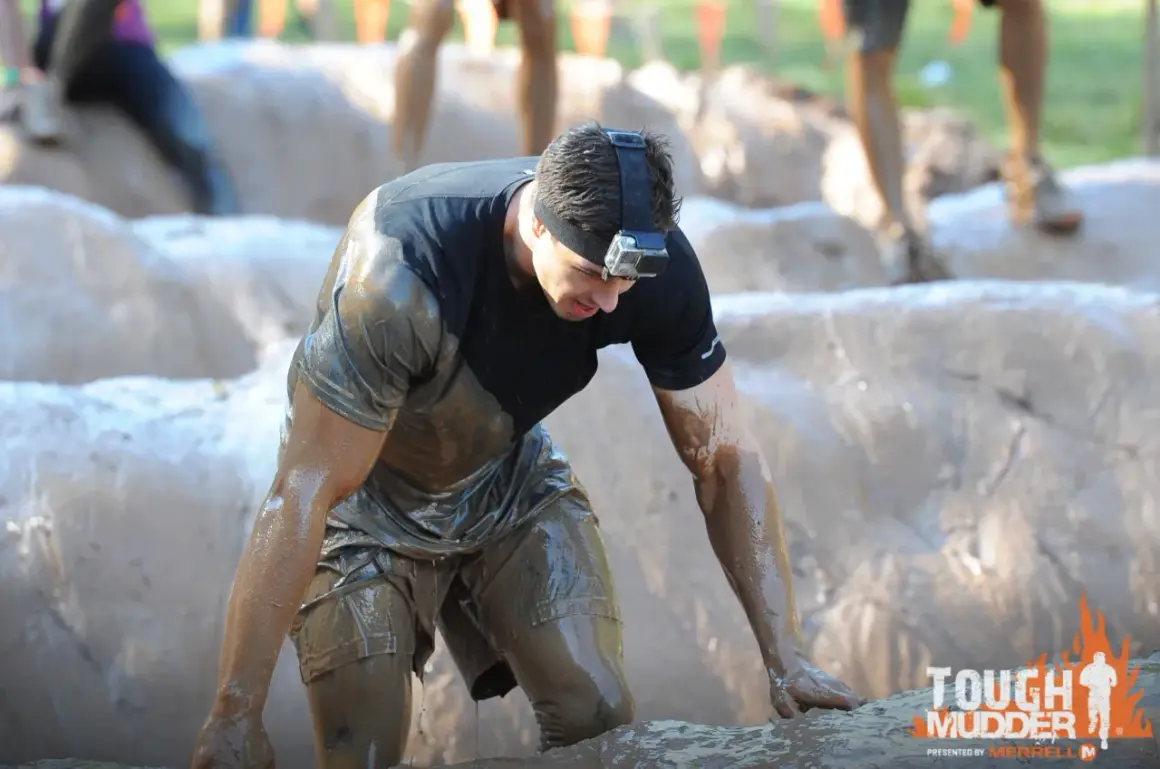 Ein Mann kämpft sich durch den Schlamm bei einem Tough Mudder **obstacle run**. Er trägt eine GoPro am Kopf.