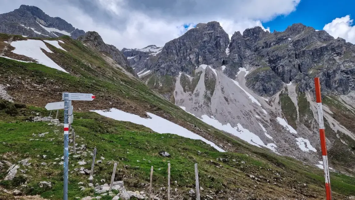 Wanderwegweiser am Widderstein Trail. Grüne Hänge mit Schneefeldern und steile Felsgipfel im Hintergrund.