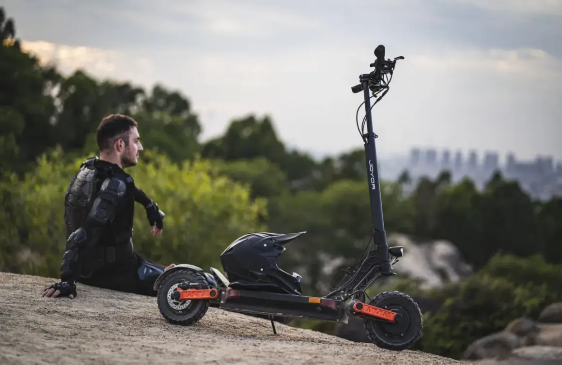 Mann in Schutzkleidung sitzt neben E-Roller mit Helm. Der e roller preis pro minute ist hier nicht ersichtlich, aber die Fahrt verspricht Abenteuer.