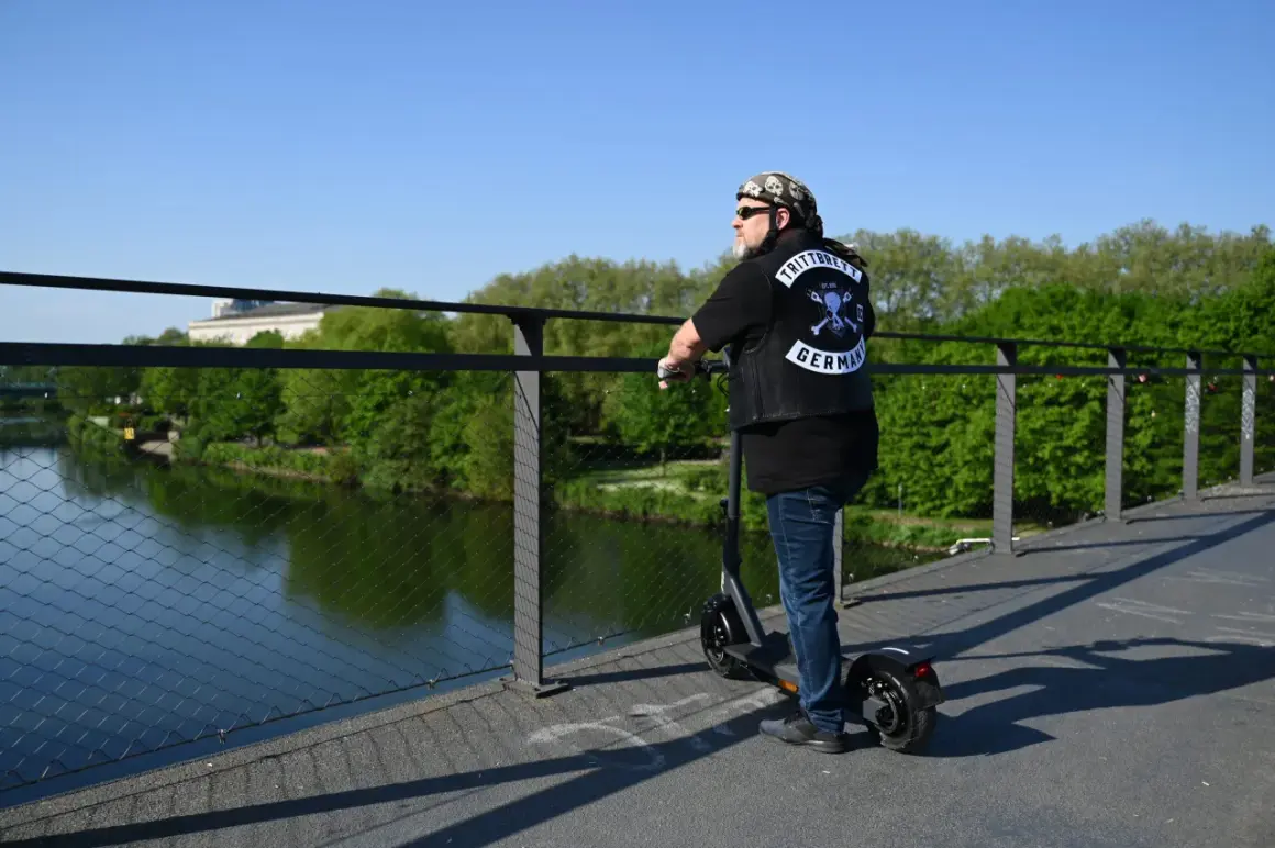 Mann auf E-Scooter mit "TRITTBRETT GERMANY" Aufnäher auf Weste. Er erklärt, wie funktioniert ein E-Scooter, während er auf einer Brücke steht.