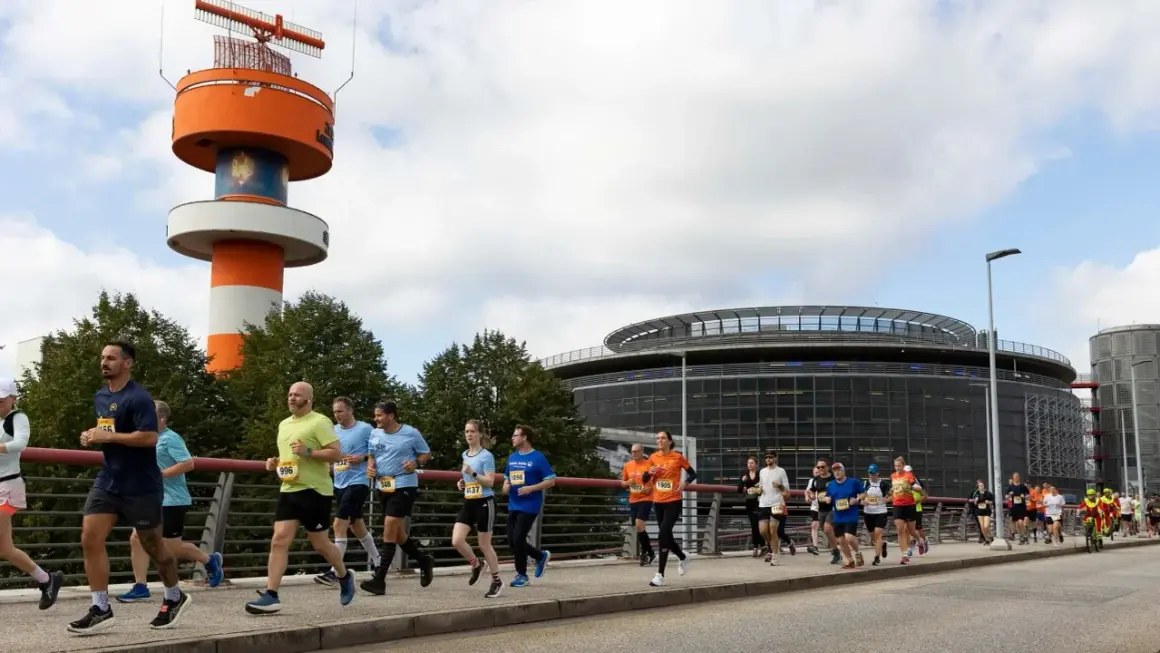 Läufer bei der Airport Race Hamburg, vorbei an einem markanten orange-weißen Turm und einem modernen Gebäude.